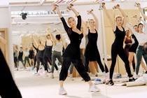 photo of reflection in mirror of Anderson leading a fitness class with both arms raised above head and hands holding weights in well-lit studio