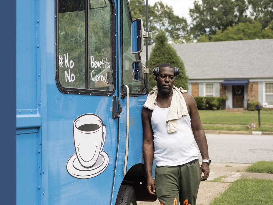 man in white tank top leaning against a blue food truck in a drive way with houses behind