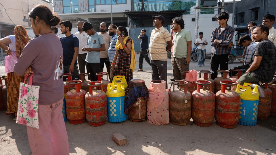 Indians wait in a long line beside red, pink, and blue gas cylinders. Some are talking on the phone, while others stare ahead with their arms crossed.
