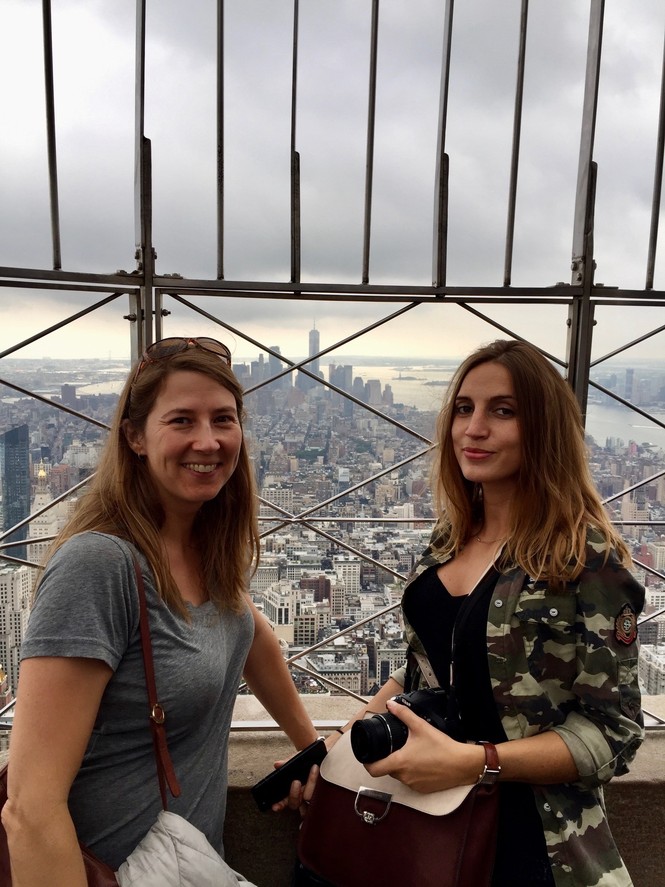Two women smiling in front of the New York City skyline at the top of a tall building.