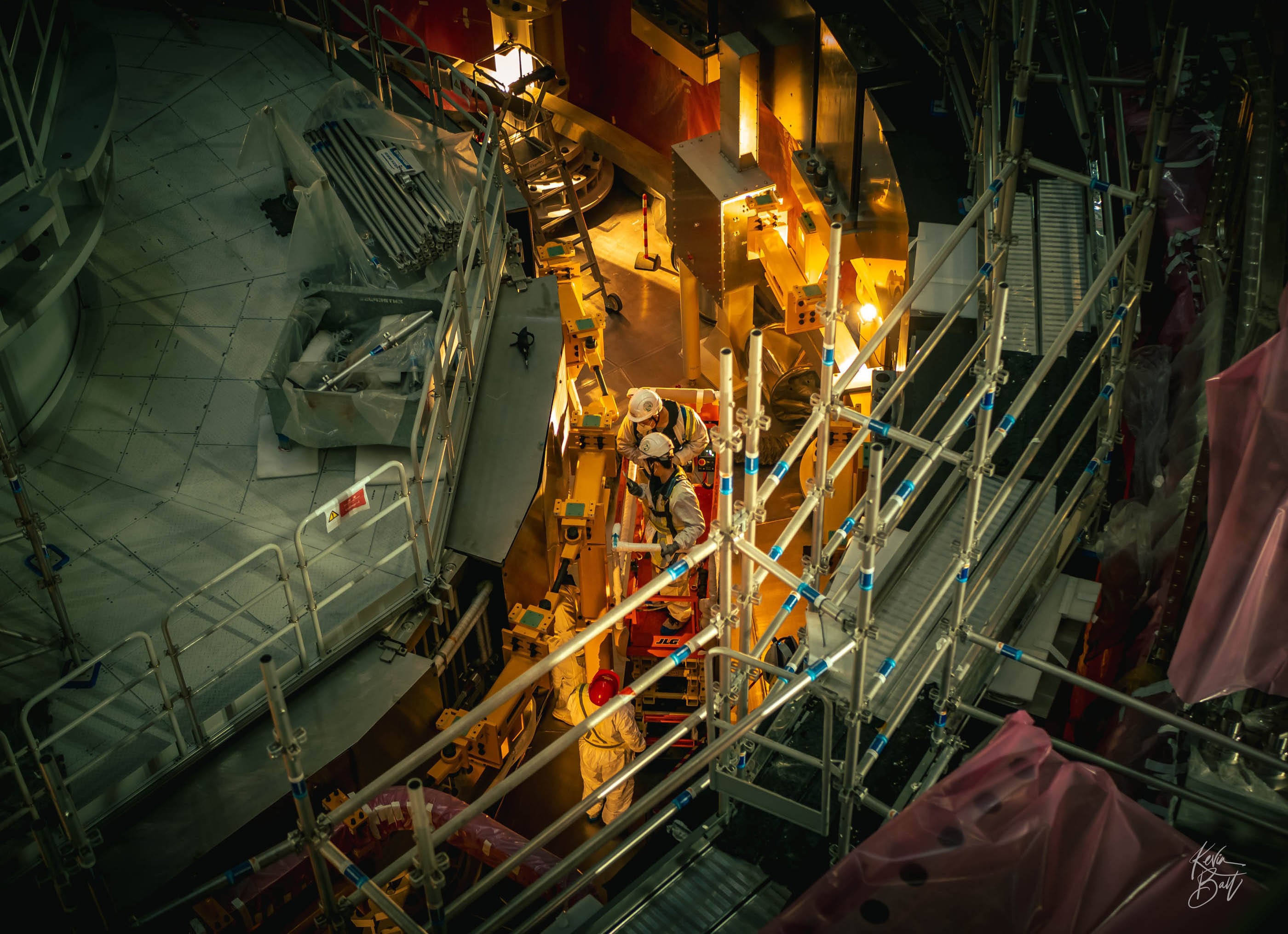 Several workers stand in and around a scissor lift near components of a fusion reactor.