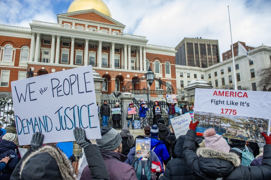 Protesters stand on the steps outside a capitol building, holding signs that read 