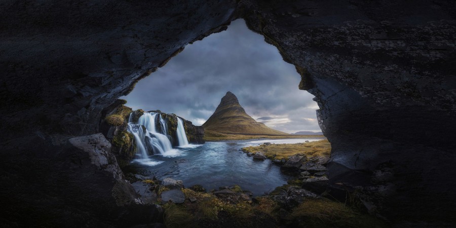 A view of a lone conical mountain in Iceland, framed by the opening of a cave, beside a waterfall