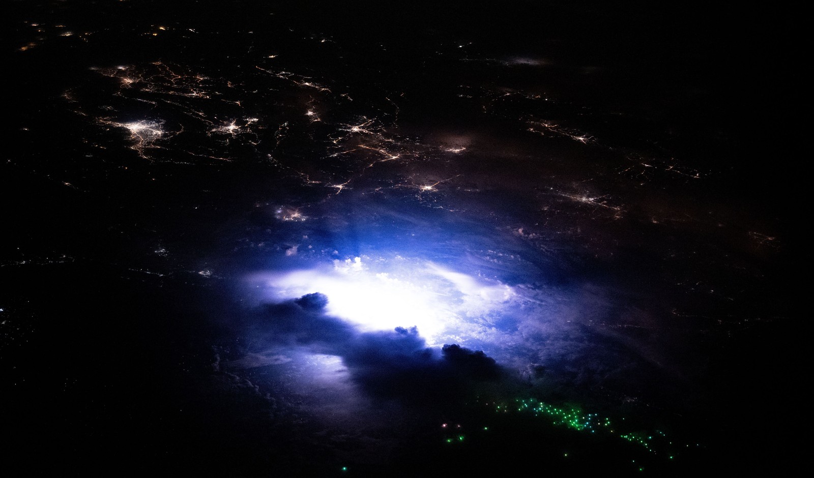 Lightning strikes in a small storm cloud near city lights, seen from orbit.