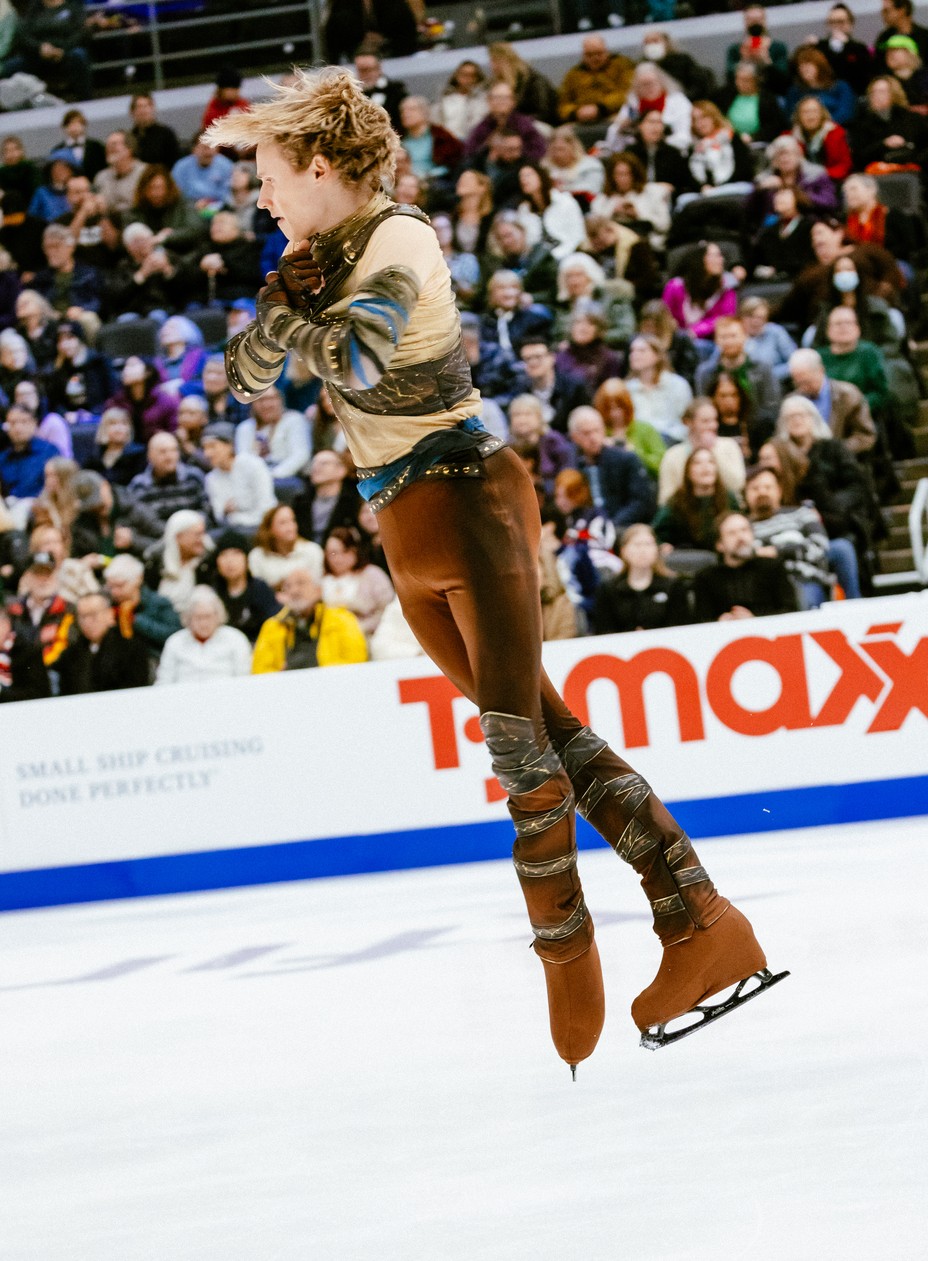 photo of young man with red hair in brown skating costume mid spin with large crowd in background