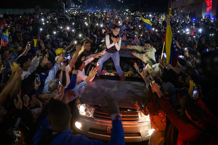 A crowd of political supporters cheer and greet a person who sits on top of a car passing through the crowd.