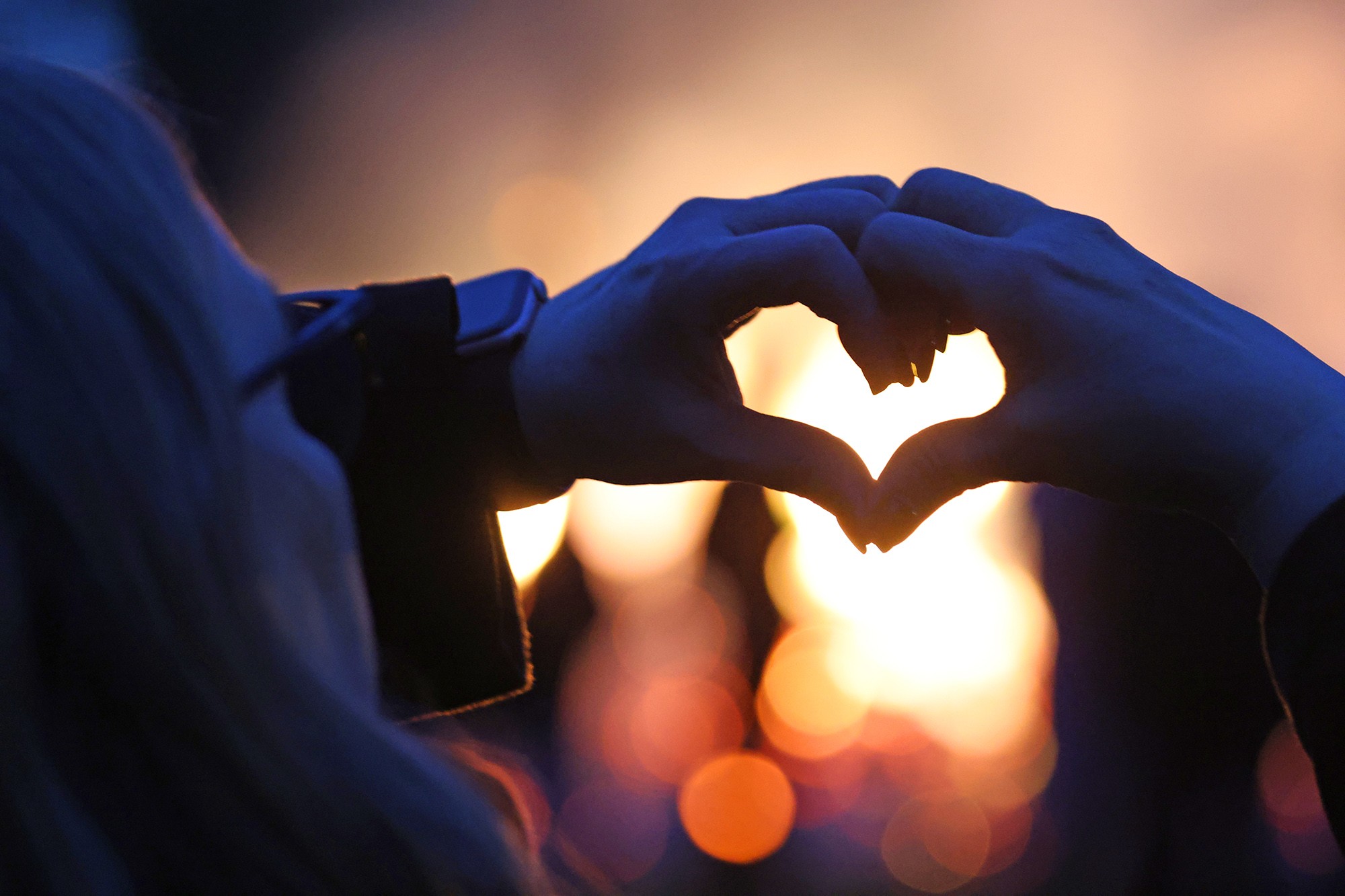 A woman stands in front of a bonfire, forming a heart with her hands.