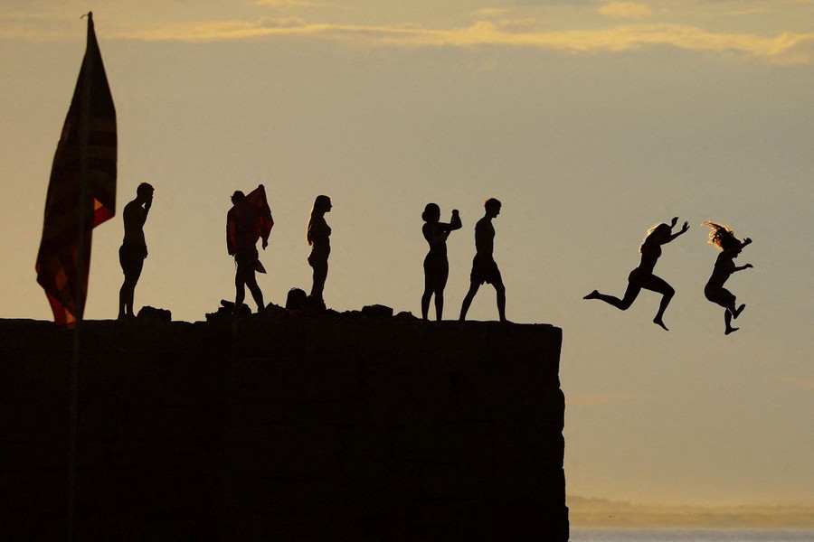 Kids jump off a breakwater into the ocean, seen in silhouette.