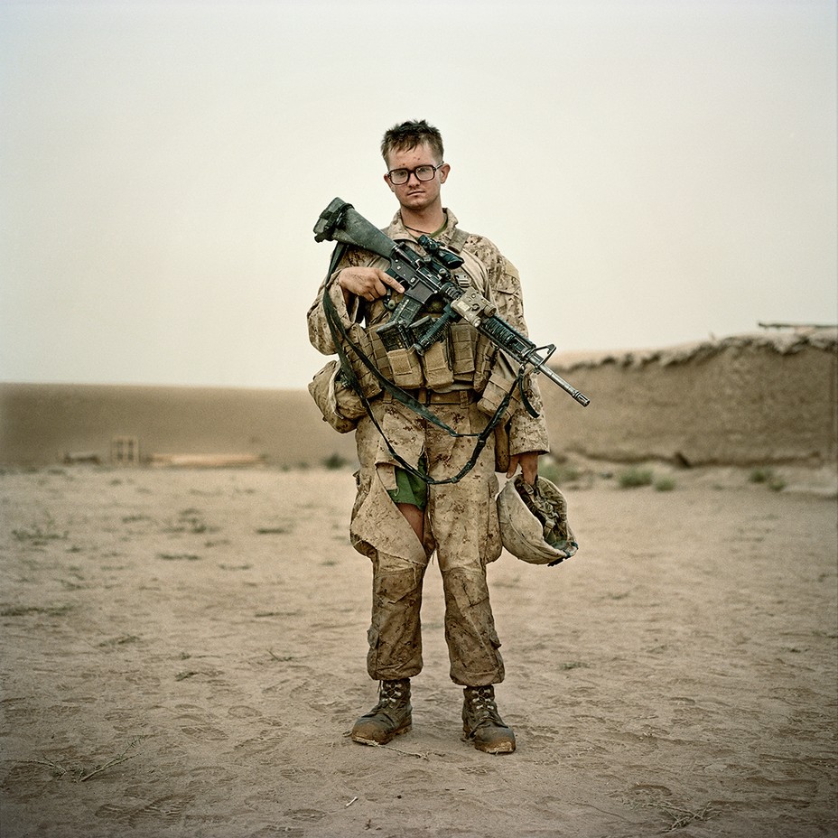Marine wearing glasses geared up and holding rifle and helmet, pants torn