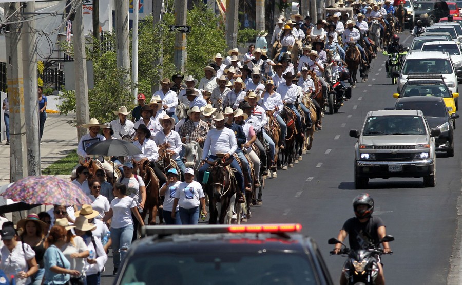 A long line of riders on horses travels slowly down a main street.