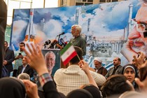 A white-haired man stands at a lectern, speaking to a crowd of people in front of a mural featuring his face.