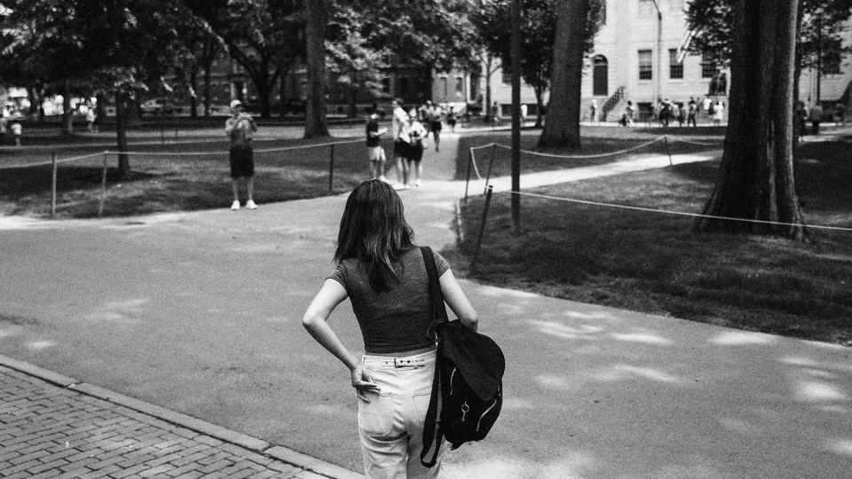 A black-and-white photo of a young woman, seen from behind, wearing a backpack and walking on a campus