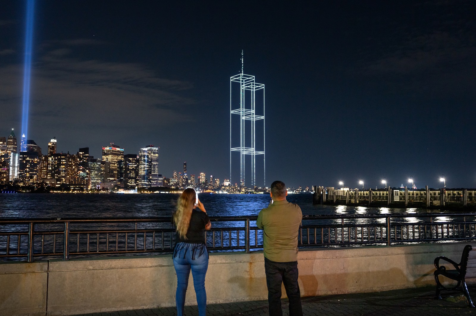 Two people take pictures as a drone light show depicting the twin towers appears alongside the New York City skyline.