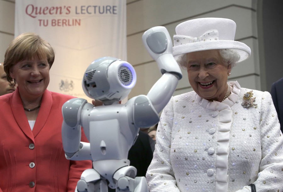 Angela Merkel and Queen Elizabeth smile as a little robot waves to the Queen.