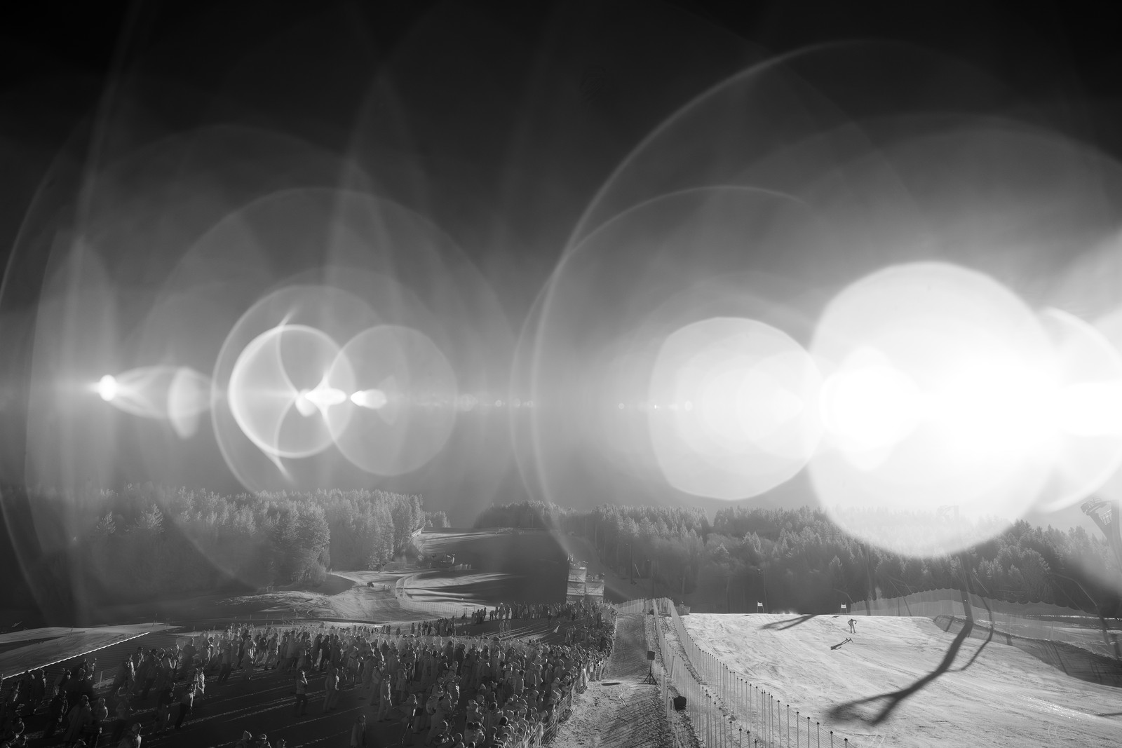 Chaotic lens flares fill the sky above a crowd watching a skier race past.