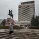 Picture of a woman wearing winter rain gear and standing up in the stairs at a square with an equestrian statue and pine trees. In the background, there is a brutalist building with a sign that reads "Cosmos."