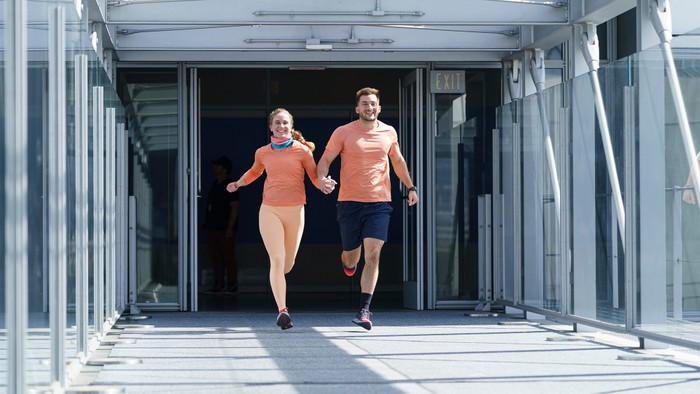 A still from the TV show The Amazing Race featuring a couple team in light orange athletic clothing running through an airport terminal