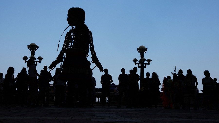 A tall marionette in silhouette stands near a crowd of people.