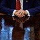 Photograph of Trump from the neck down with his hands on his desk in he