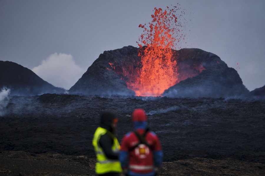 Two people stand, watching lava err, erupt from a nearby volcanic cone.