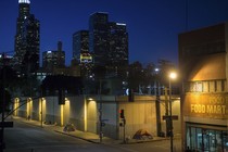 Homeless people sleeping in tents under streetlamps with the LA skyline beyond.