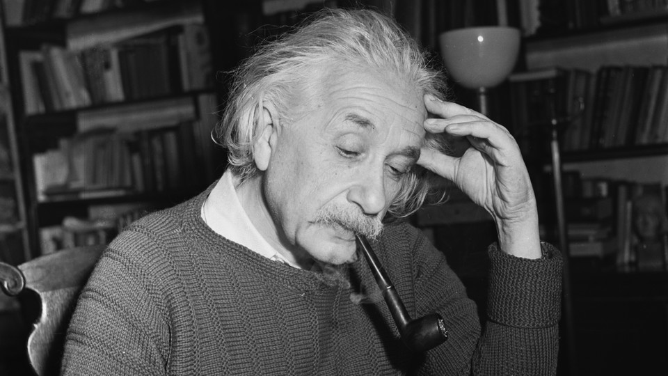 A black and white photograph of Albert Einstein at a desk, pipe in mouth, left hand against his temple