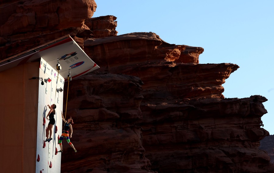 Two rock climbers compete on a man-made rock-climbing wall set up outside, beside natural cliffs.