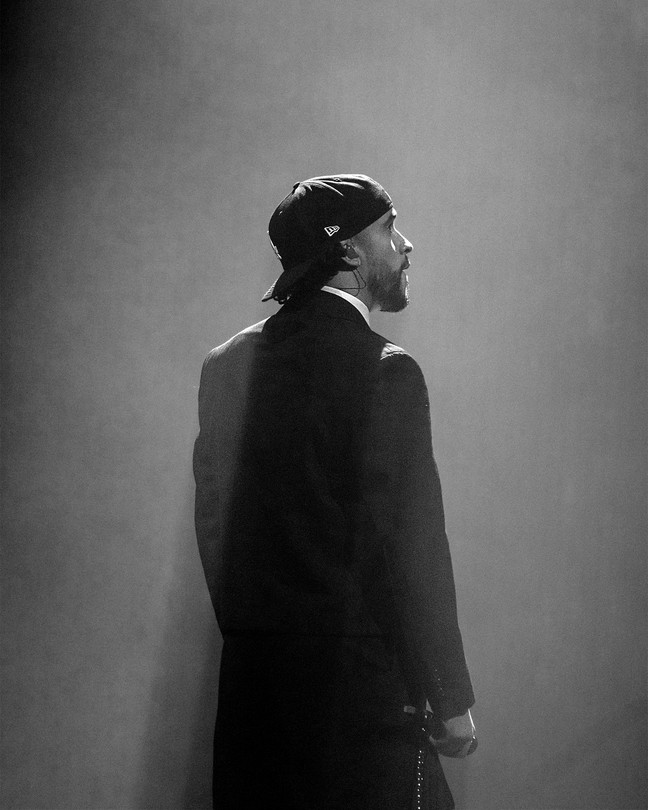 A black-and-white photo of Bad Bunny standing alone onstage in a suit and backward cap