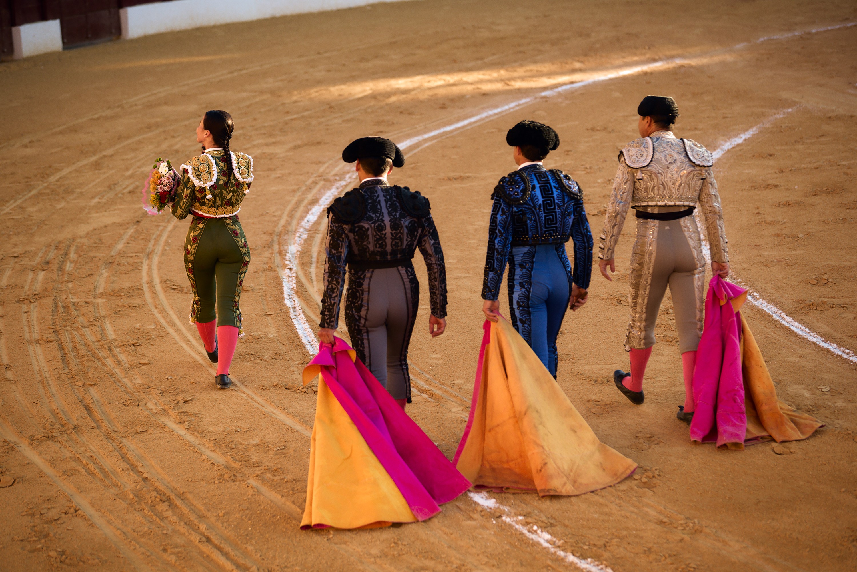 photo of woman with braid wearing ornate bullfighting uniform walking away in ring from viewer, carrying bouquet and waving to crowd, trailed by 3 men also in ornate uniforms and dragging large magenta and gold capes.