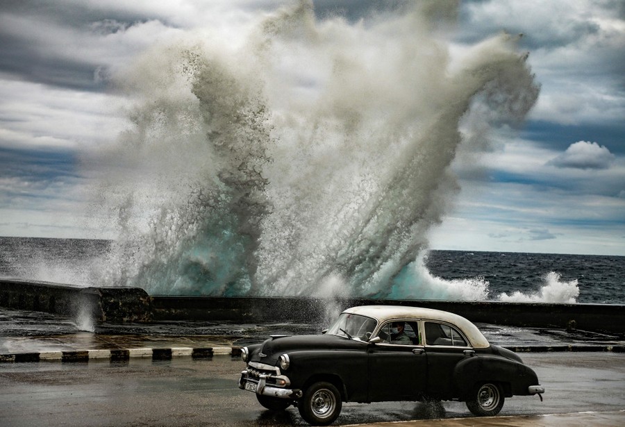 A vintage car drives past a crashing wave.