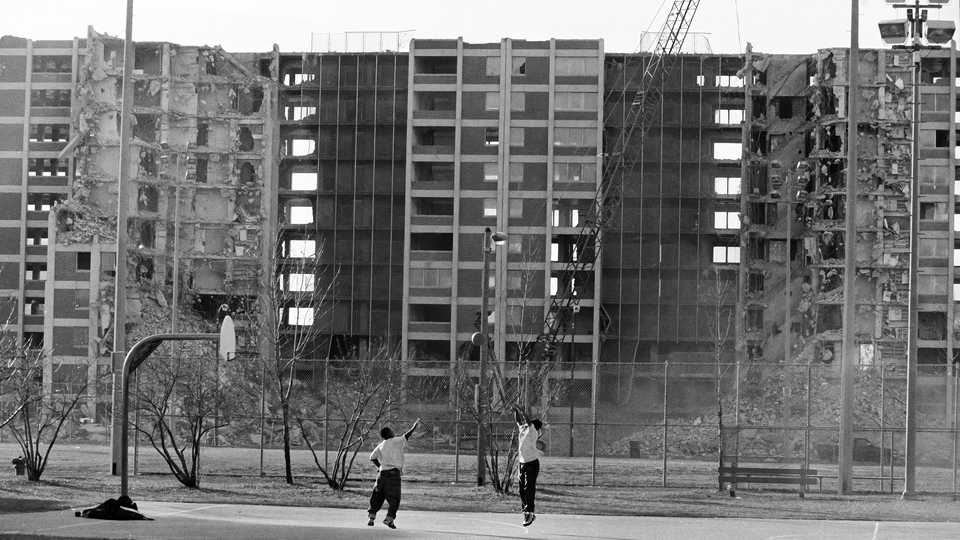 A black and white photograph of two figures playing basketball in front of a ruined housing project