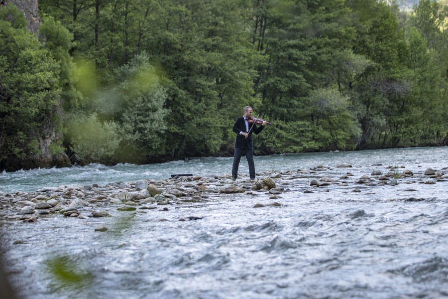 A person wearing a suit jacket plays a violin while standing in a shallow area in a rushing river.