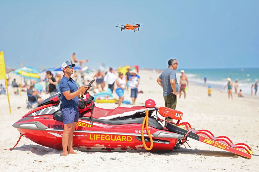A lifeguard operates a small drone on a beach, near people enjoying a warm day.