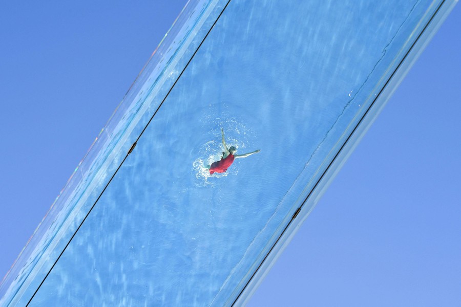 A model swims in a transparent swimming pool, high above the photographer.