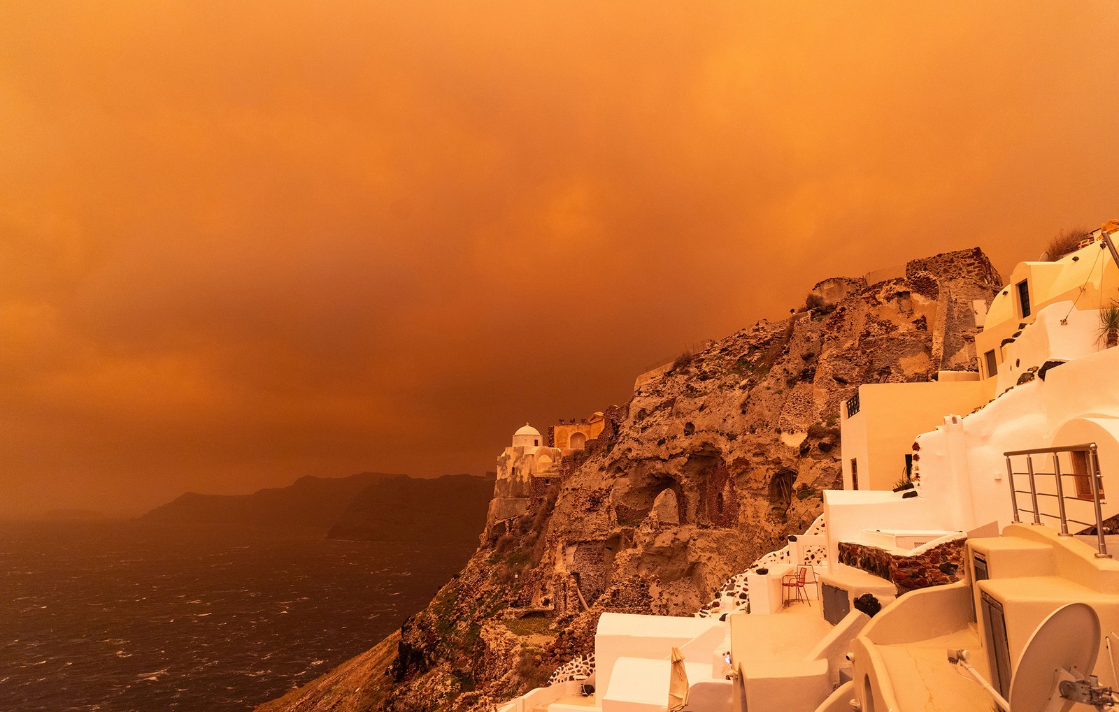 Dark, orange colored clouds, seen from the steep coast of a Greek island with many white buildings.