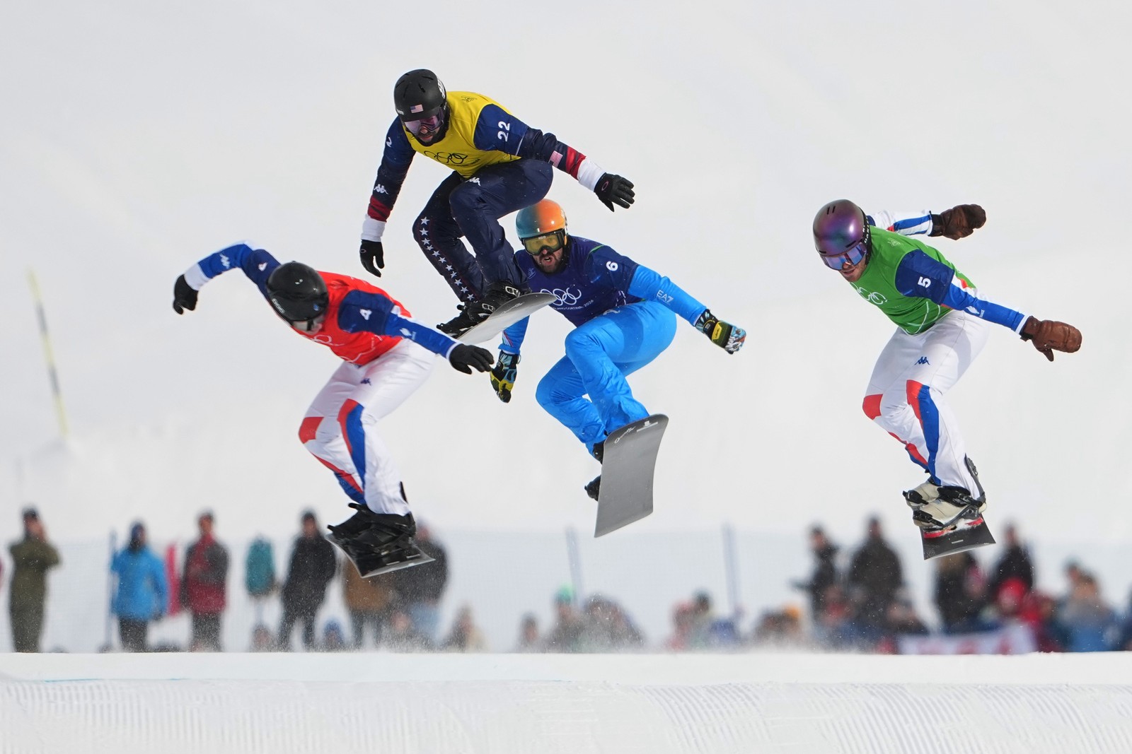 Four snowboarders make a jump as they race down a snow-covered hill.