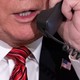 A close-up shot of Donald Trump wearing a black suit jacket, a white shirt, and a red-and-black-striped tie, holding a black phone receiver in front of his mouth