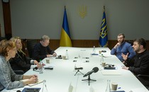 Several people sit around a large table framed by two blue-and-yellow flags; Volodymyr Zelensky speaks while several others take notes.