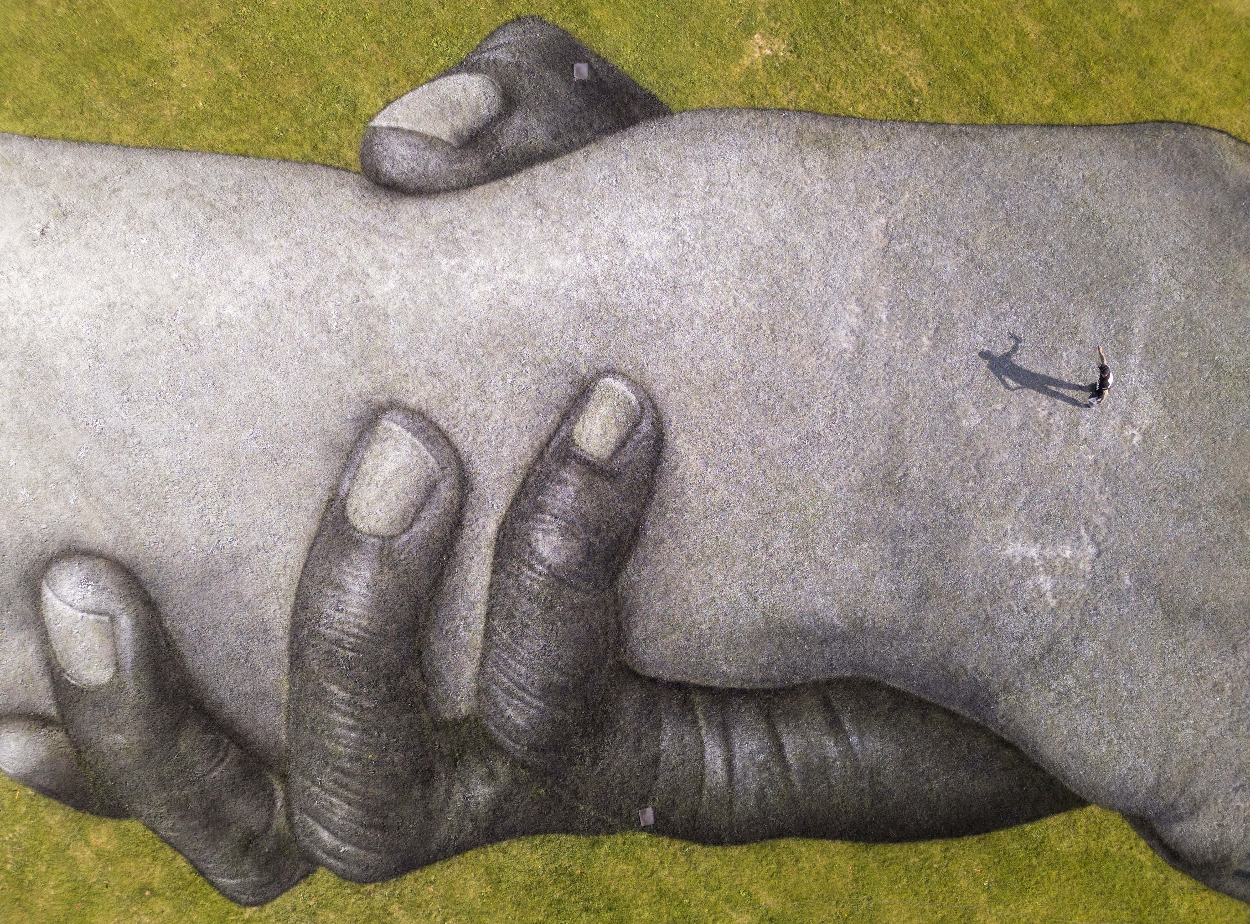 A drone view of mural painted across an expanse of grass in a park, depicting two people's arms grasping each other