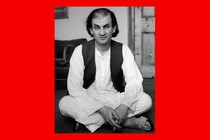 In a black-and-white image from 1988, Salman Rushdie, with chin-length dark hair and wearing a dark vest over white shirt and pants, sits crossed-legged on the floor.