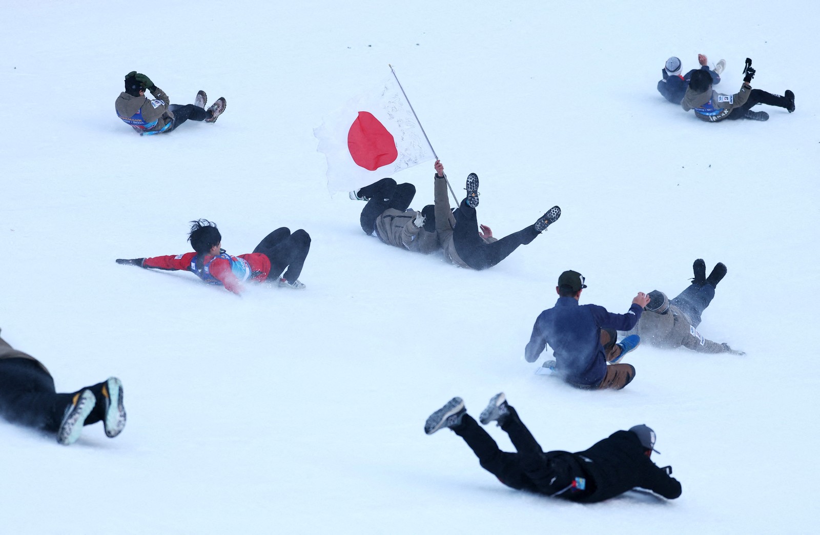 About 10 people slide and tumble down a steep snow-covered hill.