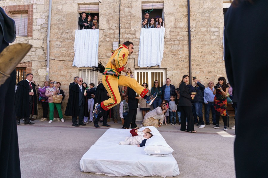 A person in costume makes a running leap over a mattress with two babies lying on it, in a street in a Spanish village.