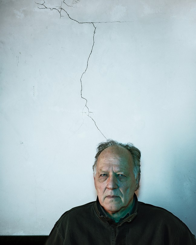 Photograph of Werner Herzog staring intently at the camera. The gray wall behind him has a large crack in it.