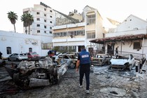 A person wearing a blue PRESS vest and carrying a large camera walking into a burned-out parking lot in Gaza.