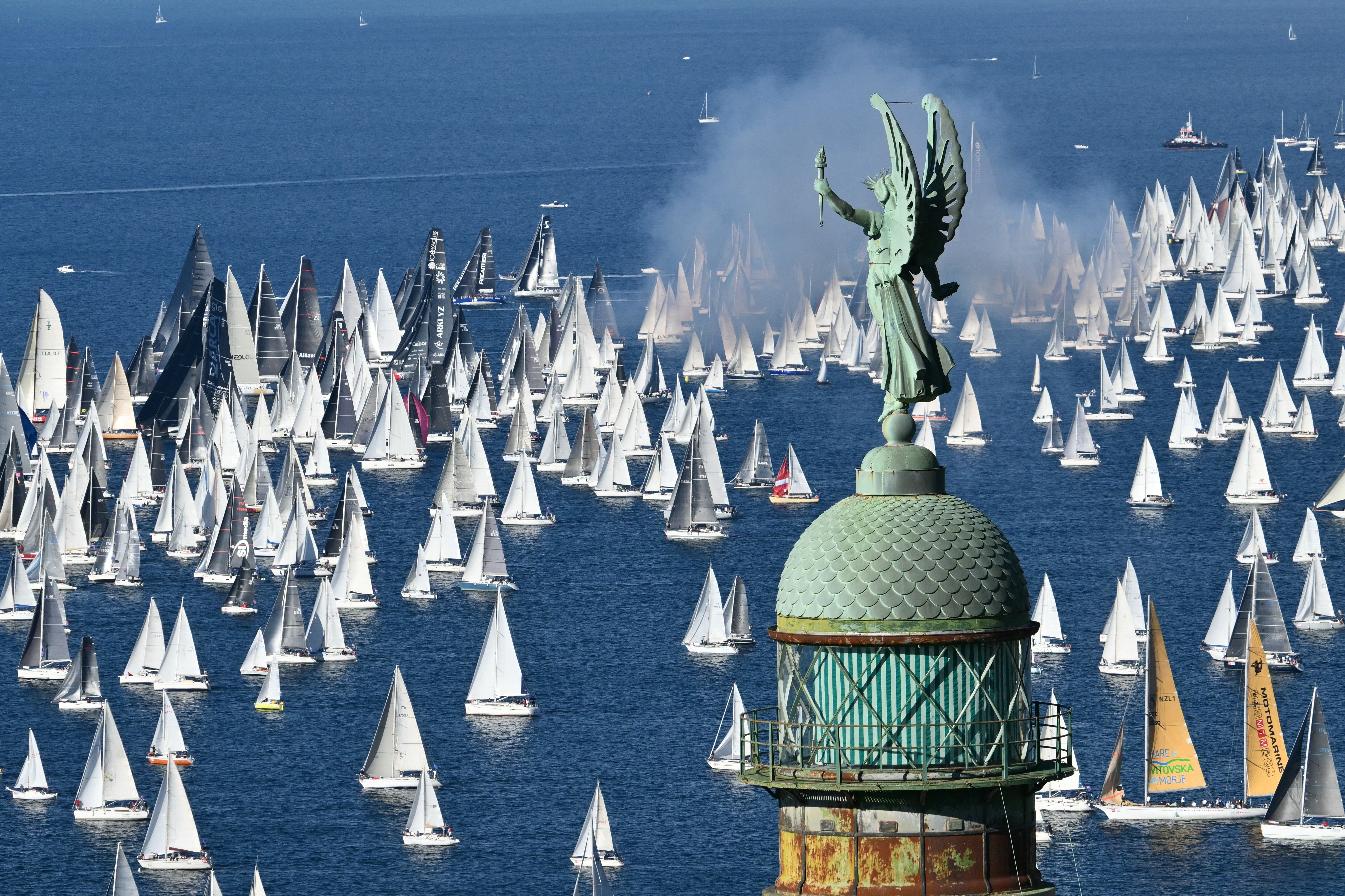 Dozens of sailboats sail together in a harbor, seen behind an ornate lighthouse.
