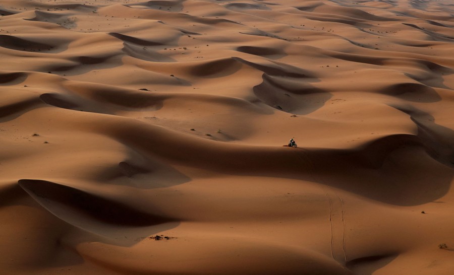 A motorcyclist rides over dunes in a broad desert.