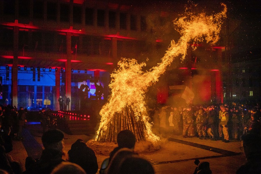 A crowd gathers around a bonfire in a city square.
