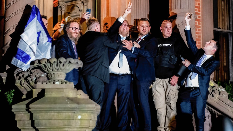 A color photograph showing Israeli National Security Minister Itamar Ben-Gvir visiting the Yale University John C. Anderson House; he holds up a "V" hand signal, for "Victory," as security guards attempt to hold him back and people hold up their cellphones to take pictures.
