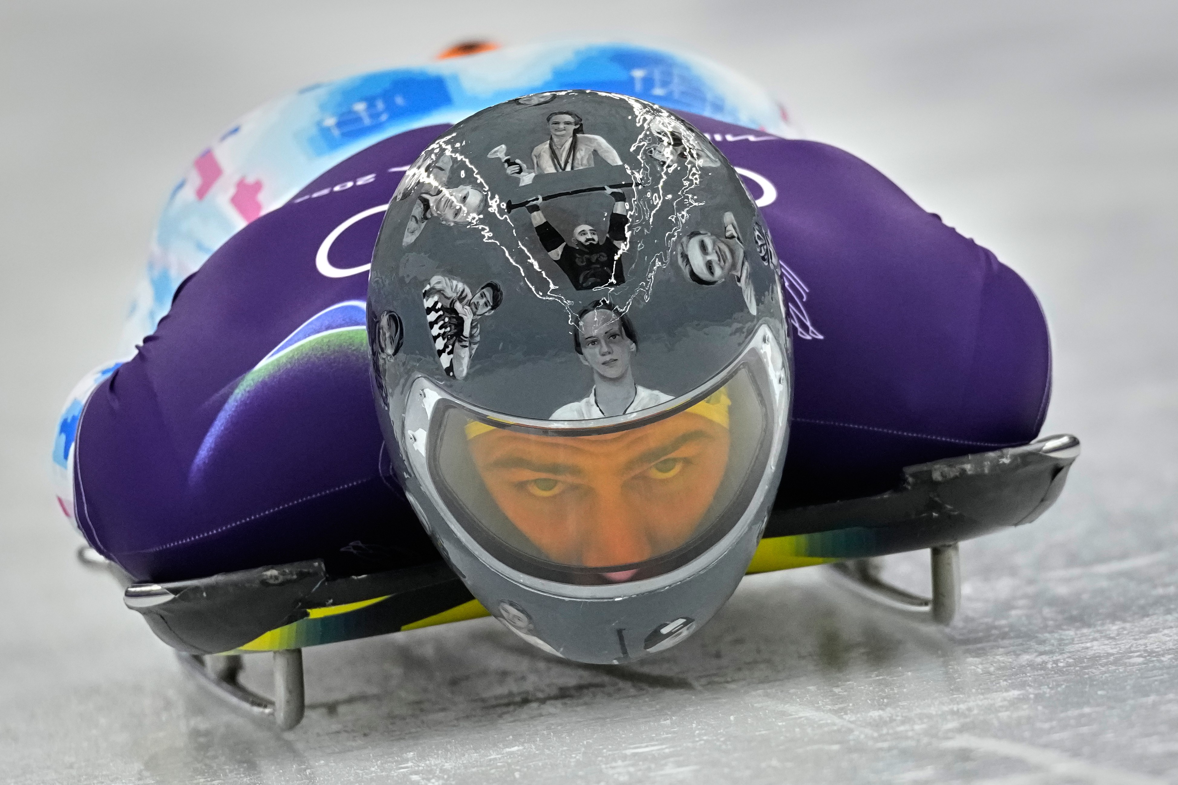 An athlete lies flat on a luge sled during a run, looking up. Their helmet is adorned with many images of other athletes. 