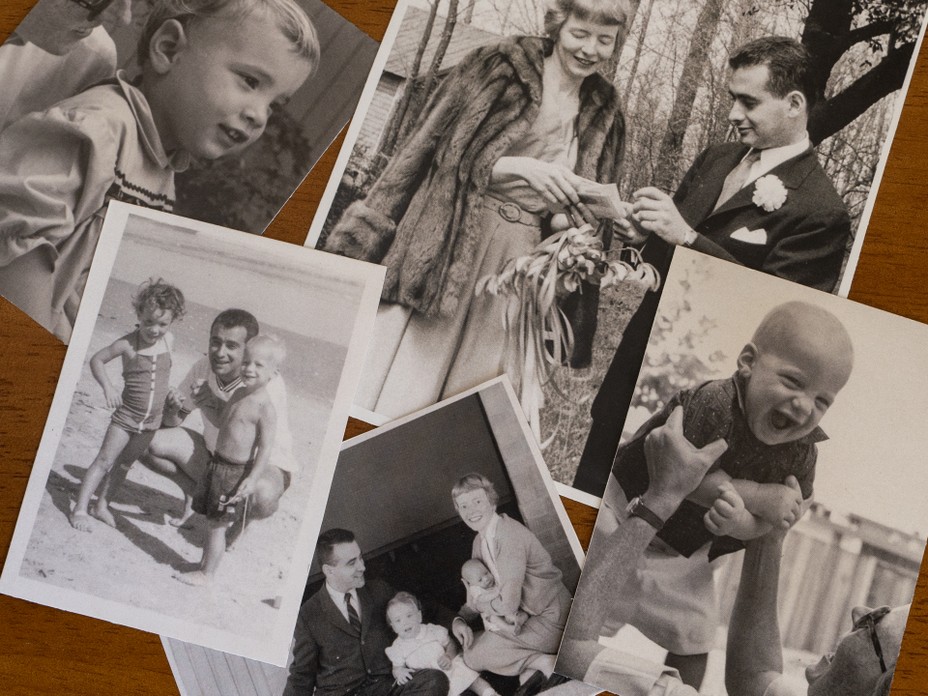 photo of an array of black-and-white family photos arranged on a wooden table, including portrait of child, two children at beach with man, wedding photo, family photo of couple with baby and toddler, and man lifting baby